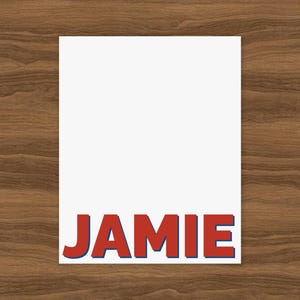 May include: A white rectangular card with the name "JAMIE" printed in bold, red letters with a blue shadow at the bottom. The card is set against a brown wooden background.