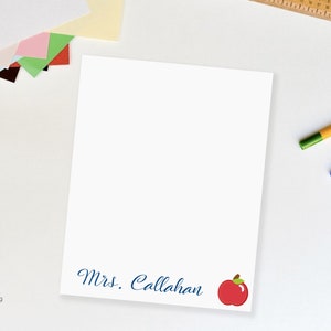 Może przedstawiać: Biały notatnik z napisem "Mrs. Callahan" w niebieskim skrypcie, z grafiką czerwonego jabłka. Ołówki i kolorowy papier są widoczne w tle. Notatnik jest prawdopodobnie dla nauczyciela.