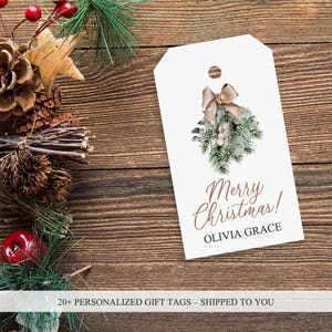 Pode incluir: Etiqueta de presente branca com uma ilustração em aquarela de um ramo de vegetação com um laço marrom. O texto "Merry Christmas!" está escrito em letras cursivas marrons. O nome "Olivia Grace" está escrito em letras pretas abaixo da saudação.