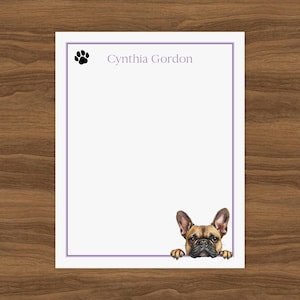 以下が含まれることがあります： 紫色の縁取り、足跡、上部に「Cynthia Gordon」という名前が入った白い文房具。フレンチブルドッグのイラストが下から覗いています。文房具は木製の表面にあります。