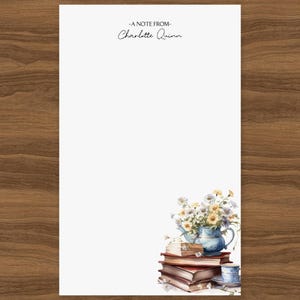 May include: White stationery with a watercolor illustration of a stack of books, a blue teapot filled with flowers, and a teacup. The text "-A NOTE FROM- Charlotte Quinn" is printed at the top. The stationery is on a wooden surface.