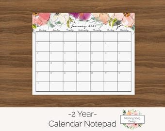 Può includere: Un blocco note calendario mensile per gennaio 2027 con un bordo floreale ad acquerello nei toni del rosa, viola e verde nella parte superiore. La griglia include i giorni della settimana e le date numerate. Sotto, il testo dice 2 Year Calendar Notepad.