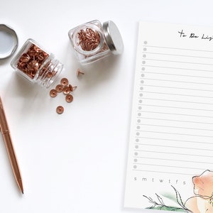 May include: A white to-do list notepad with a floral design and the words "To Do List" at the top. The notepad has a column for each day of the week. There are two jars of rose gold push pins and paper clips on a white surface.