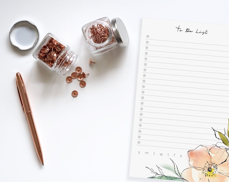 May include: A white to-do list notepad with a floral design and the words "To Do List" at the top. The notepad has a column for each day of the week. There are two jars of rose gold push pins and paper clips on a white surface.