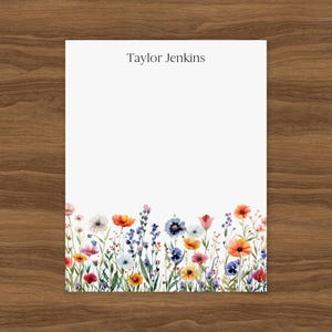 Könnte beinhalten: Weißes Briefpapier mit einem Aquarell-Blumenrand mit bunten Wildblumen in Rot-, Orange-, Blau- und Lilatönen. Der Text "Taylor Jenkins" ist oben aufgedruckt. Das Briefpapier wird auf einer Holzoberfläche präsentiert.