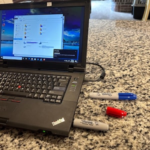 May include: A black laptop computer with a ThinkPad logo on the bottom left corner. The laptop is open and the screen is displaying a Windows desktop. There are two blue and gray Sharpie markers and a red marker on the surface.