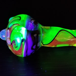 Może przedstawiać: Szklana fajka w kolorze zielonym, fioletowym i czerwonym z wzorem w postaci spirali. Fajka świeci pod czarnym światłem.