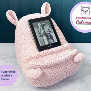 May include: A pink crocheted Kindle Paperwhite holder with ears. The holder is 11.7 inches wide and 16.9 inches tall. The text "Creator of Cuteness" is visible in a purple circle with a pink llama illustration.