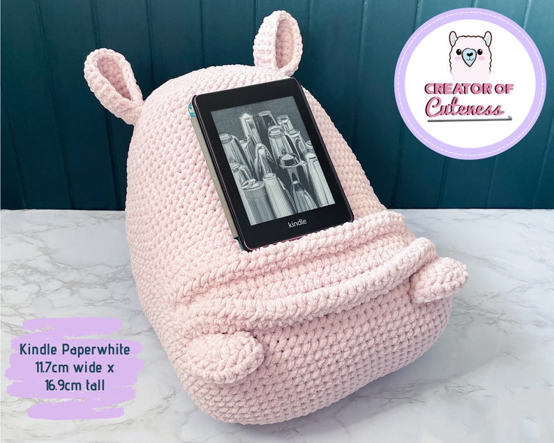 May include: A pink crocheted Kindle Paperwhite holder with ears. The holder is 11.7 inches wide and 16.9 inches tall. The text "Creator of Cuteness" is visible in a purple circle with a pink llama illustration.