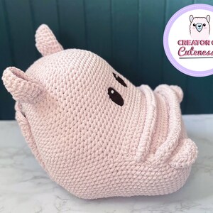 May include: A pink crocheted hippopotamus stuffed animal with black eyes. The hippopotamus is lying on its side with its head turned to the right. The hippopotamus is made of a soft, plush material.