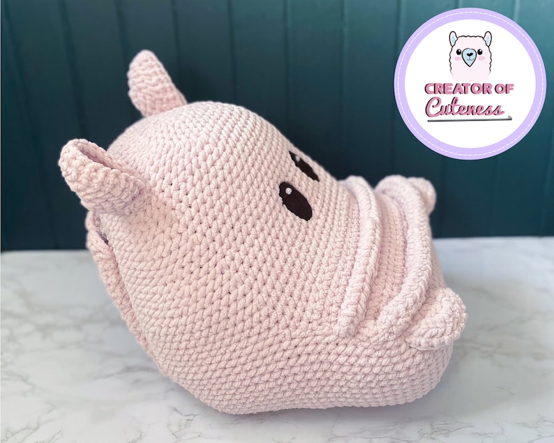 May include: A pink crocheted hippopotamus stuffed animal with black eyes. The hippopotamus is lying on its side with its head turned to the right. The hippopotamus is made of a soft, plush material.