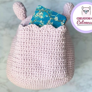 May include: A pink crocheted stuffed animal with a pocket on the front. The pocket has a blue and white patterned book inside. The stuffed animal has two small ears on top.