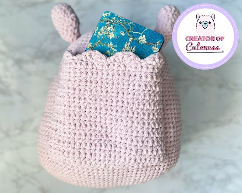 May include: A pink crocheted stuffed animal with a pocket on the front. The pocket has a blue and white patterned book inside. The stuffed animal has two small ears on top.