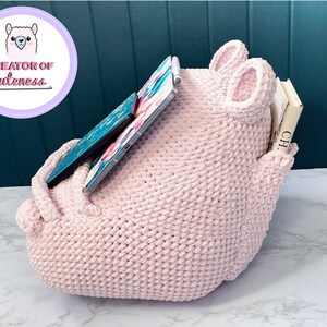 May include: A pink crocheted storage organizer with two large ears, a pocket on the side, and a tablet and books inside.