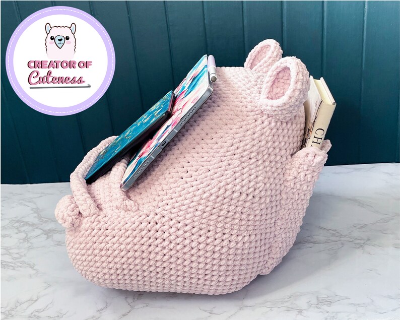 May include: A pink crocheted storage organizer with two large ears, a pocket on the side, and a tablet and books inside.