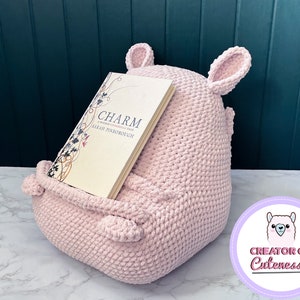 May include: A pink crocheted book holder shaped like a llama with large ears. The book holder is holding a book titled "Charm: A Woman's Chronicle" by Sarah Pinborough. The book holder is sitting on a white marble surface. The text "Creator of Cuteness" is on a purple circle with a white llama illustration.
