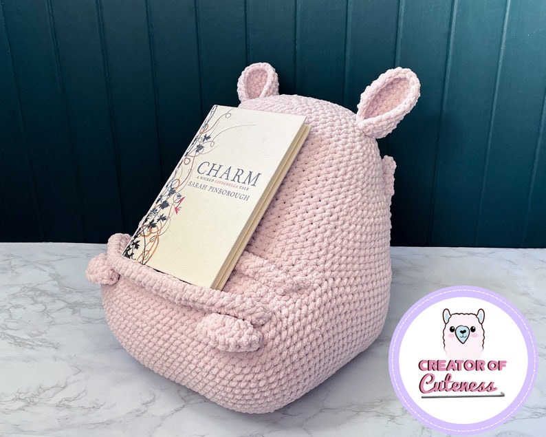 May include: A pink crocheted book holder shaped like a llama with large ears. The book holder is holding a book titled "Charm: A Woman's Chronicle" by Sarah Pinborough. The book holder is sitting on a white marble surface. The text "Creator of Cuteness" is on a purple circle with a white llama illustration.