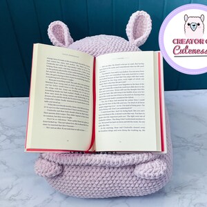 May include: A light purple crocheted book holder shaped like a llama with large ears. The book holder is holding a book open with a red bookmark. The text on the book reads "It was almost as if she didn't even go to sleep. But having  a book in her hand, she remembered what life was like  before she met the prince."