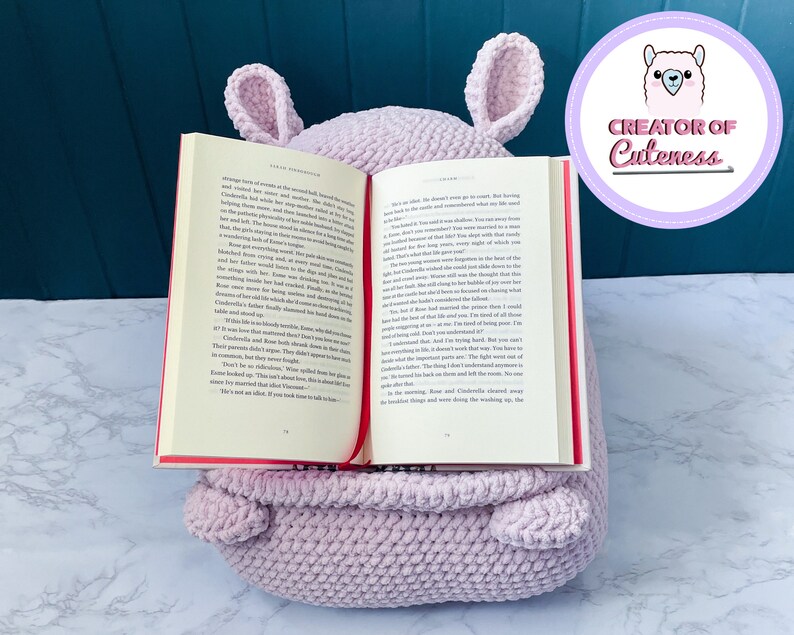 May include: A light purple crocheted book holder shaped like a llama with large ears. The book holder is holding a book open with a red bookmark. The text on the book reads "It was almost as if she didn't even go to sleep. But having  a book in her hand, she remembered what life was like  before she met the prince."
