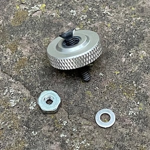 Puede incluir: Un tornillo de metal plateado con cabeza moleteada, una tuerca de metal plateado y una arandela de metal plateado. El tornillo está roscado y tiene una punta negra. La tuerca es hexagonal y tiene un agujero en el centro. La arandela es redonda y tiene un agujero en el centro.