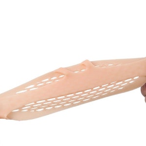 May include: A beige, flexible foot care product with a honeycomb pattern and finger separators is stretched between two hands. The product appears to be made of a soft, pliable material, designed for foot comfort and support.