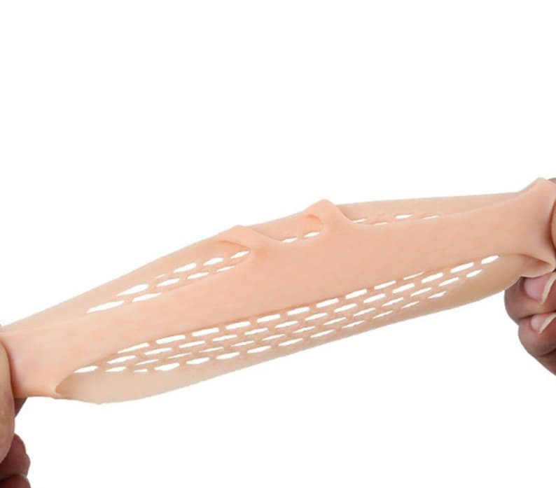 May include: A beige, flexible foot care product with a honeycomb pattern and finger separators is stretched between two hands. The product appears to be made of a soft, pliable material, designed for foot comfort and support.