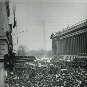 May include: A black and white photograph of a large crowd gathered in front of a building. A man is suspended upside down from a crane, with the text "HOUDINI SUSPENDED IN STRAIGHT JACKET WASHINGTON JAN 12 1922" written on the bottom of the image.
