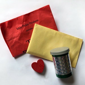 May include: A red envelope with the text "Ickle Pickle Products - presents" on it, a yellow envelope, a red heart, and a green and silver tube with a grey lid.