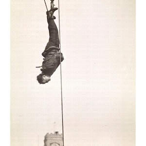 May include: A man is suspended upside down from a rope attached to a pulley system. He is wearing a dark jacket and trousers. A clock tower can be seen in the background.