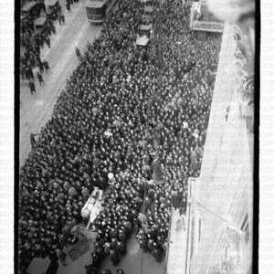 May include: Aerial view of a large crowd of people gathered in a city street. A horse-drawn carriage is visible, along with streetcars and other vehicles. The image is in black and white.