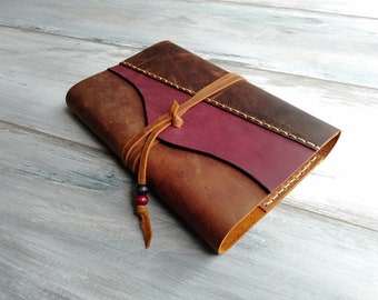 May include: A brown leather journal with a burgundy leather flap and a brown leather strap. The journal has a decorative bead tassel.