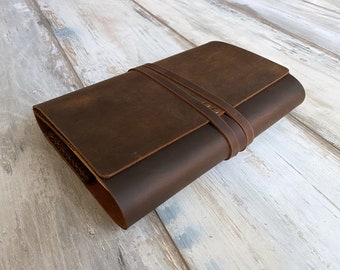 May include: A brown leather journal with a strap closure. The journal is closed and lying on a light wood surface.