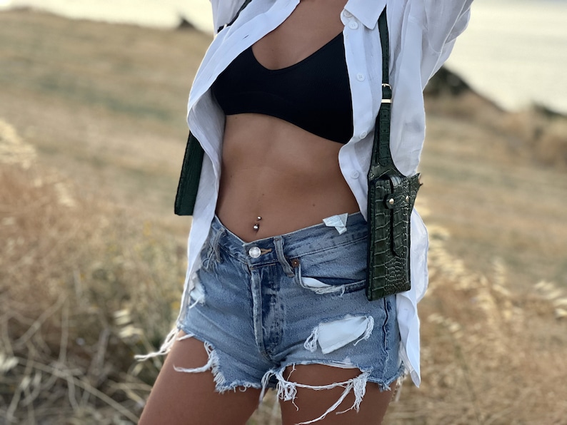 May include: A woman wearing a white button-down shirt, a black bra, and denim shorts. She is carrying a green crocodile-embossed leather crossbody bag.