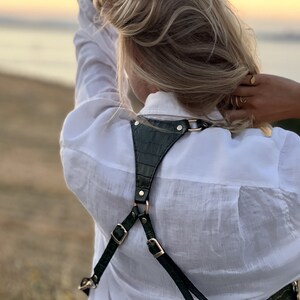 May include: A green leather camera strap with gold hardware. The strap is worn over the shoulders and has a criss-cross design at the back.