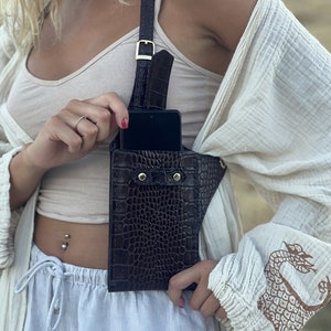 May include: A brown leather crossbody phone bag with a crocodile pattern. The bag has a strap that goes over the shoulder and a flap that closes with a snap. The bag is worn by a person with a white top and white trousers.