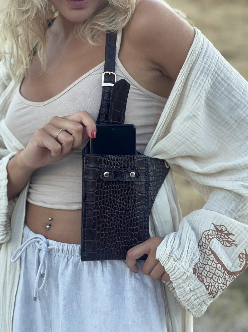 May include: A brown leather crossbody phone bag with a crocodile pattern. The bag has a strap that goes over the shoulder and a flap that closes with a snap. The bag is worn by a person with a white top and white trousers.