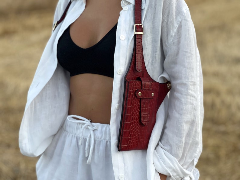 May include: A red leather crossbody bag with a crocodile-embossed design. The bag has a strap that goes over the shoulder and a buckle closure. The bag is worn over a white shirt and white trousers.