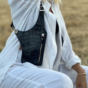 May include: A black leather crossbody bag with a gold zipper and buckle. The bag is worn over the shoulder of a person wearing a white shirt.
