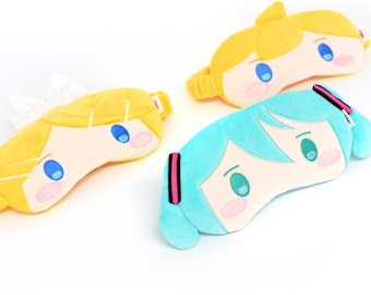 以下が含まれることがあります： アニメのキャラクターが描かれた、3つのぬいぐるみ製のアイマスク。マスクは黄色、青、水色。それぞれのマスクには、大きな目と小さな口をした漫画の顔が描かれています。