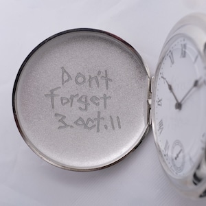 May include: A silver pocket watch with a white face and black hands. The inside of the watch case is engraved with the words "Don't Forget 3.Oct.11".