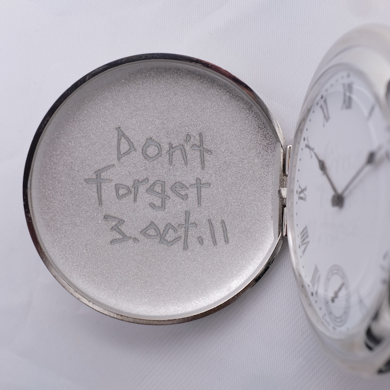 May include: A silver pocket watch with a white face and black hands. The inside of the watch case is engraved with the words "Don't Forget 3.Oct.11".