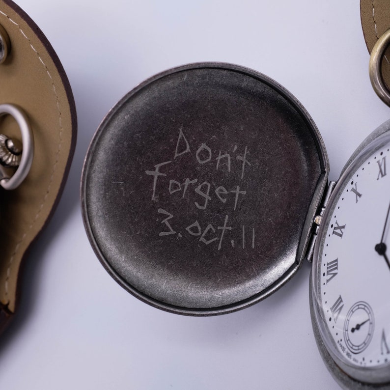 May include: A silver pocket watch with a white face and black Roman numeral hour markers. The watch is open and the inside of the case has the inscription "Don't forget 3.Oct.11" written on it.