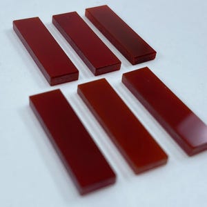 May include: Six rectangular, polished, deep red stones are arranged on a white surface. The stones are evenly spaced and have a smooth, glossy finish. The color is a rich, saturated red.