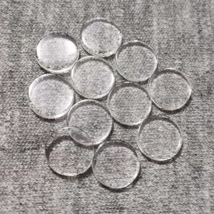 Puede incluir: Una colección de once cabujones de vidrio transparentes y circulares. Estas piezas de vidrio abovedadas con respaldo plano están dispuestas sobre una superficie gris texturizada. Los cabujones son transparentes, permitiendo el paso de la luz.