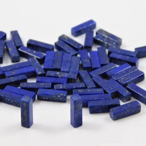 May include: A collection of small, rectangular lapis lazuli gemstones. The stones are a deep blue color with flecks of gold, and are scattered across a white surface. The gemstones are cut into a rectangular shape, and are likely used for jewelry making or other crafts.