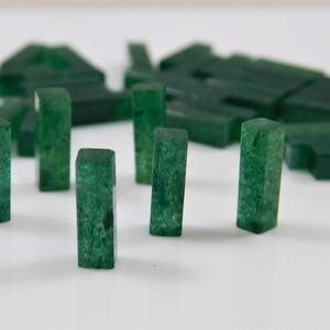 May include: A collection of small, rectangular, dark green jade stones. Some stones stand upright, while others are scattered in the background. The stones have a polished, smooth appearance and vary slightly in shape.