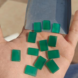 May include: A collection of rectangular, polished green gemstones. The gemstones are displayed on a hand, with a metal tray in the background. The stones have a smooth, glossy surface and a vibrant, emerald green color.