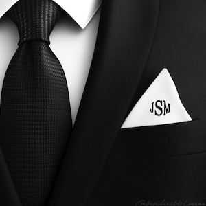 以下が含まれることがあります： 白いポケットチーフに「JSM」のイニシャルが刺繍された黒いスーツジャケット。ジャケットは、黒の模様のネクタイをつけた白いドレスシャツの上に着ています。