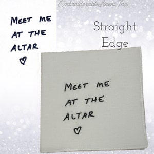 K&ouml;nnte beinhalten: Eine quadratische, cremewei&szlig;e Stoffserviette mit schwarzem Sticktext, der "MEET ME AT THE ALTAR" lautet, gefolgt von einem kleinen Herzsymbol. Die Serviette hat gerade, vern&auml;hte Kanten und ein schlichtes, klares Design f&uuml;r formelle Anl&auml;sse.