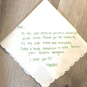 K&ouml;nnte beinhalten: Ein wei&szlig;es Taschentuch mit gewelltem Rand und gr&uuml;ner Stickerei, die folgenden Text enth&auml;lt: Dad, To dry your tears as you have always dried mine. Thank you for walking by my side today and everyday. Today a bride, tomorrow a wife, forever your favorite daughter. I love you -Maddie.